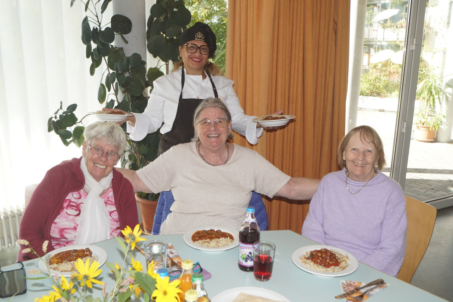 Drei sitzende Personen an einem gedeckten Tisch mit Tellern Spaghetti Bolognese, eine stehende Person mit schwarzer Schürze hält zwei Teller.