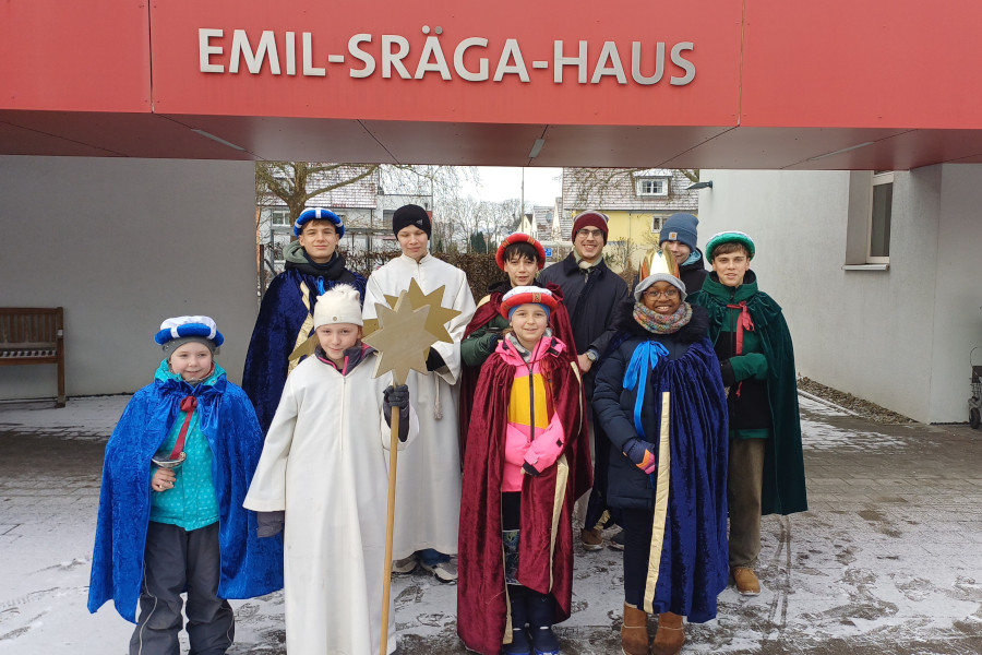 Gruppe von Kindern in bunten Umhängen und Kostümen steht vor dem Gebäude mit der Aufschrift 'EMIL-SRÄGA-HAUS'.