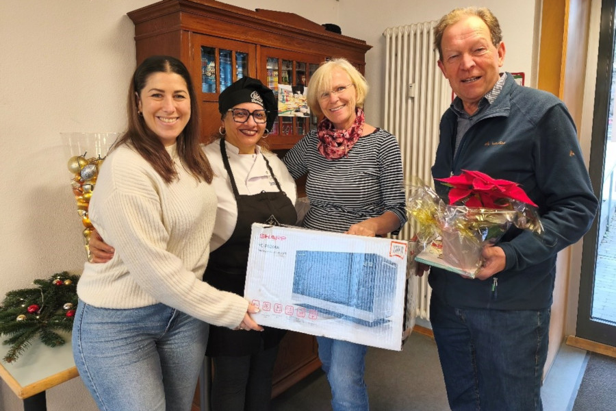 Vier Personen stehen in einem Raum vor einem Holzschrank, eine Person hält eine Mikrowellenbox, eine andere einen mit Folie verpackten Geschenkkorb mit roter Schleife.