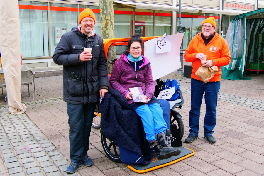 Drei Personen vor einem Stand mit AWO-Logo, eine sitzt in einem Rollstuhl, die anderen beiden stehen daneben auf gepflastertem Platz