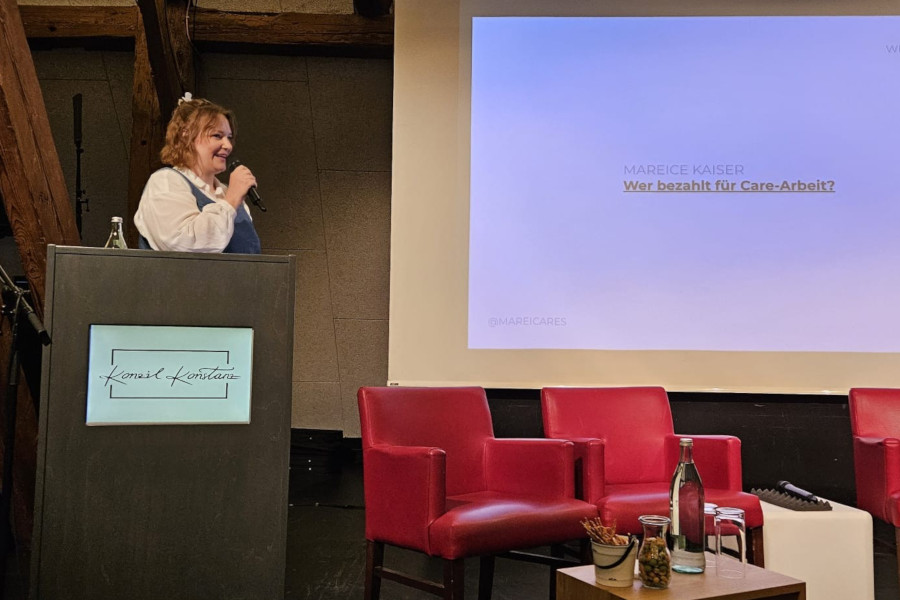 Frau mit Mikrofon steht an einem Rednerpult mit Bildschirm und spricht vor einer Leinwand mit Text, daneben drei rote Sessel und ein Tisch mit Wasserflasche und Snacks