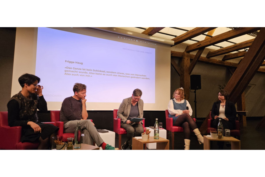 Fünf Personen sitzen auf roten Sesseln vor einer Leinwand in einem Raum mit sichtbaren Holzbalken, eine Person liest aus einem Buch