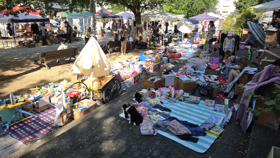 Flohmarkt mit Decken und Tischen, auf denen Kleidung, Spielzeug und andere Gegenstände zum Verkauf liegen, umgeben von Menschen und Bäumen.