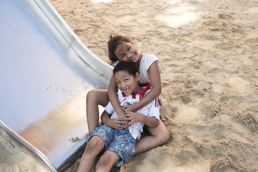 Zwei lachende Kinder sitzen im Sand am Ende einer Rutsche und umarmen sich